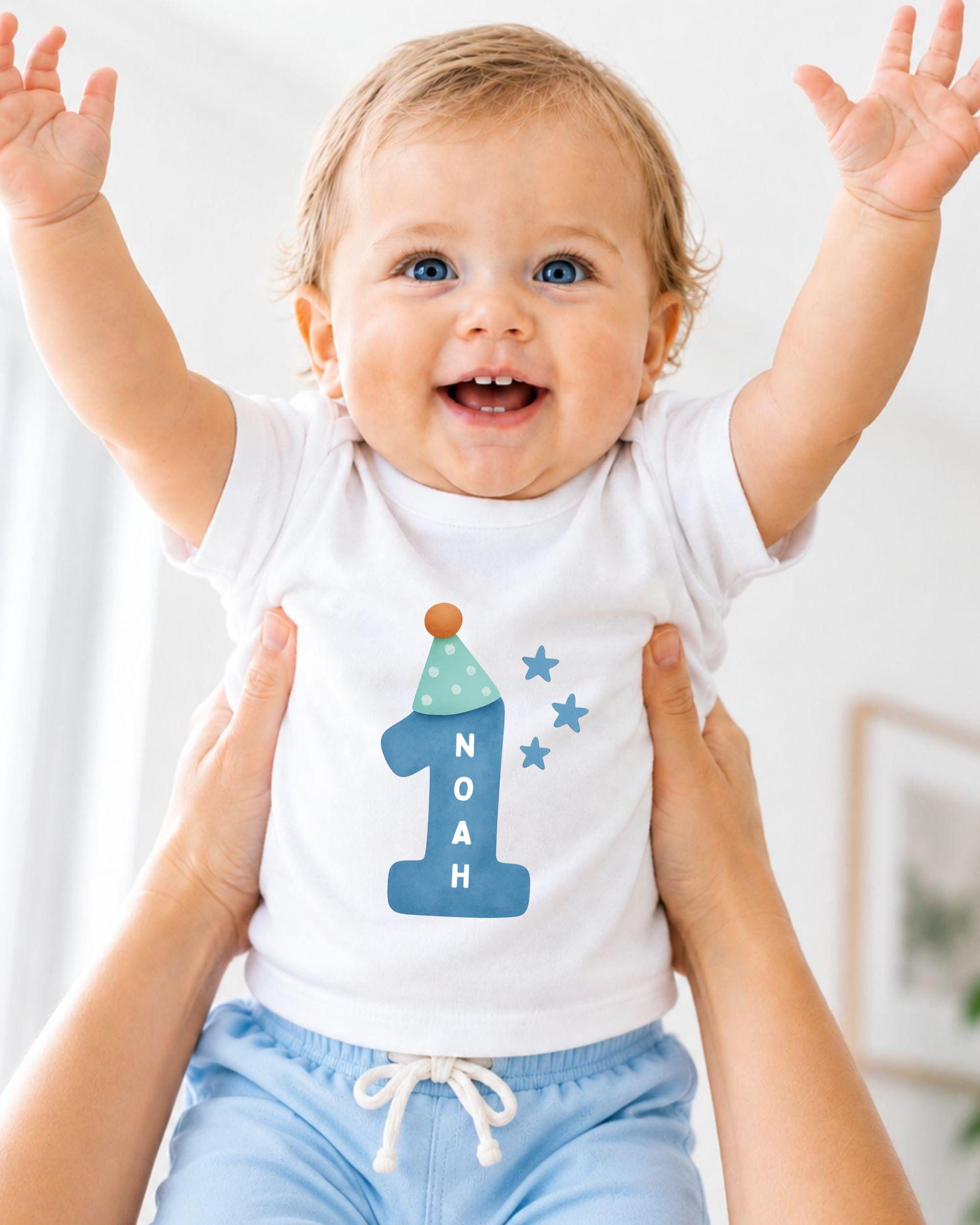 A smiling baby wearing a white onesie with a blue number one and stars, being held up by someone's arms.