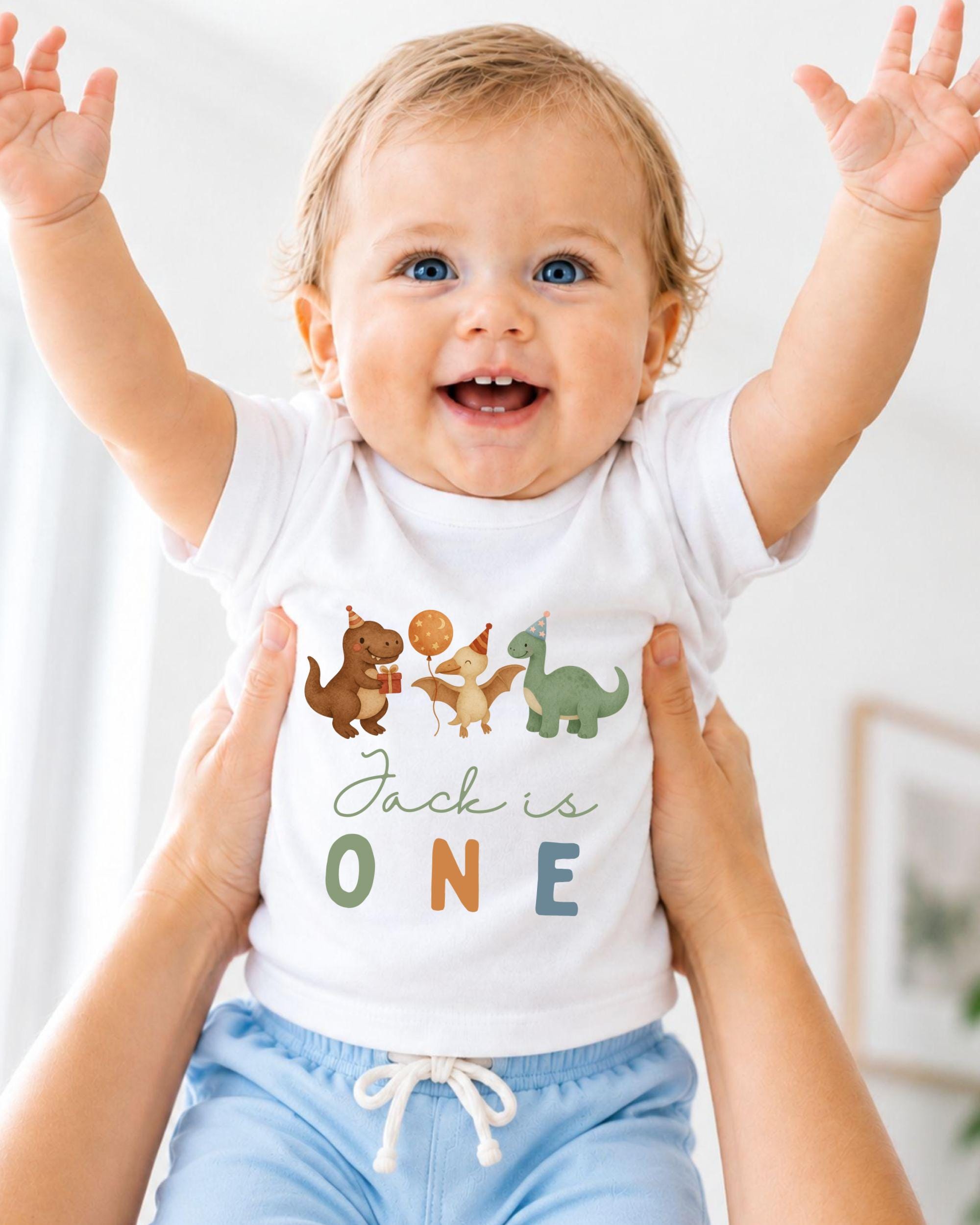 A smiling baby wearing a white t-shirt with dinosaur illustrations and the text "Jack is 1" is being held up by someone's arms.