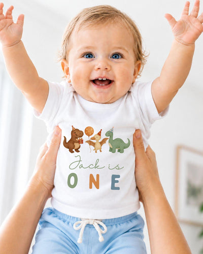 A smiling baby wearing a white t-shirt with dinosaur illustrations and the text "Jack is 1" is being held up by someone's arms.