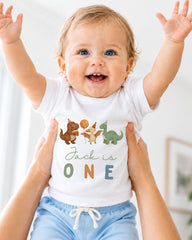 A smiling baby wearing a white t-shirt with dinosaur illustrations and the text "Jack is 1" is being held up by someone's arms.