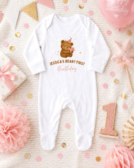 A white onesie with a teddy bear and the text " Jessica's first birthday" printed on it, surrounded by pink and gold confetti, a pink star, and a pink and gold birthday cake.