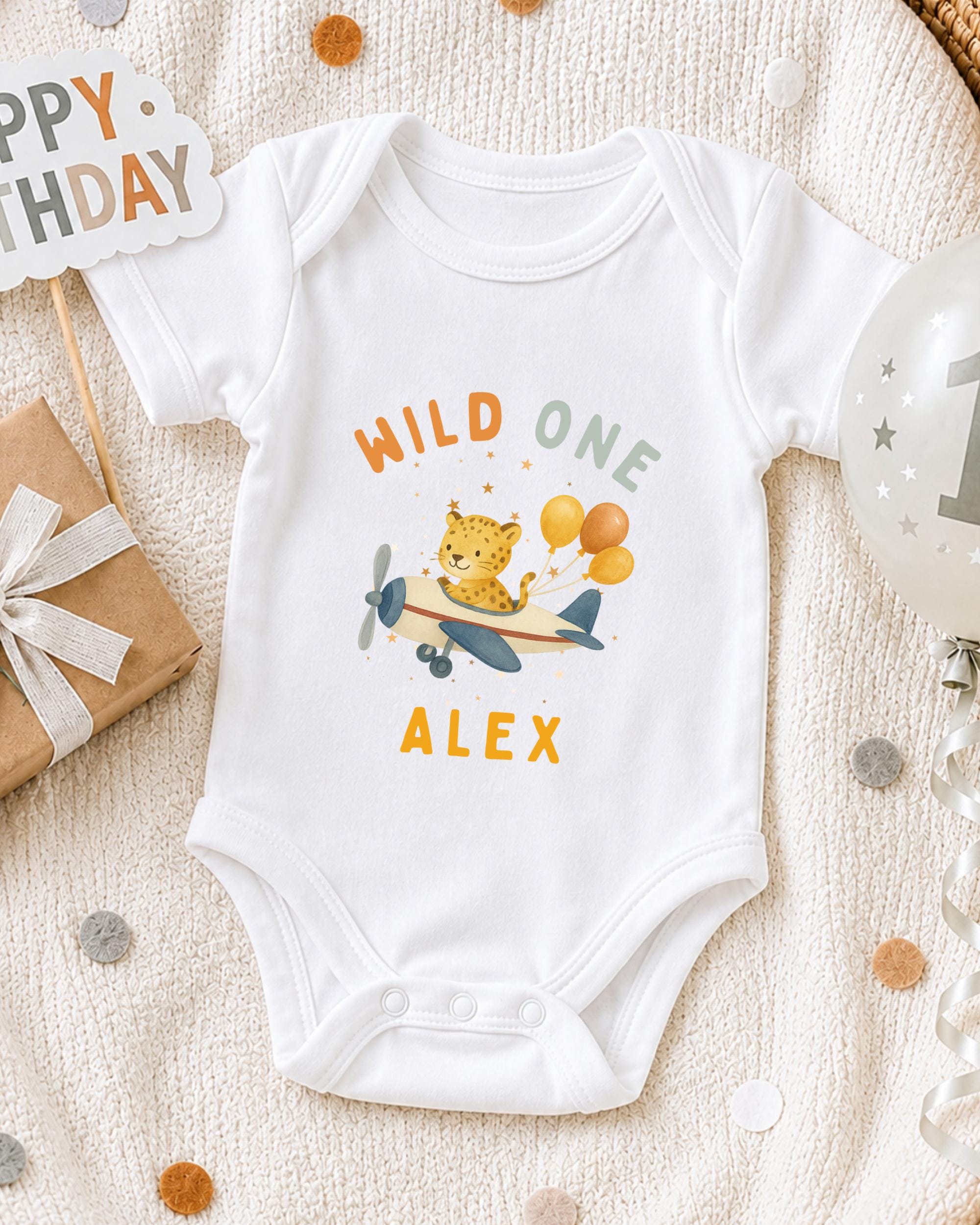A white onesie with a cartoon leopard and airplane printed on it, along with a "Happy Birthday Alex" sign and a balloon with the number "1" on it, all placed on a white blanket with confetti.
