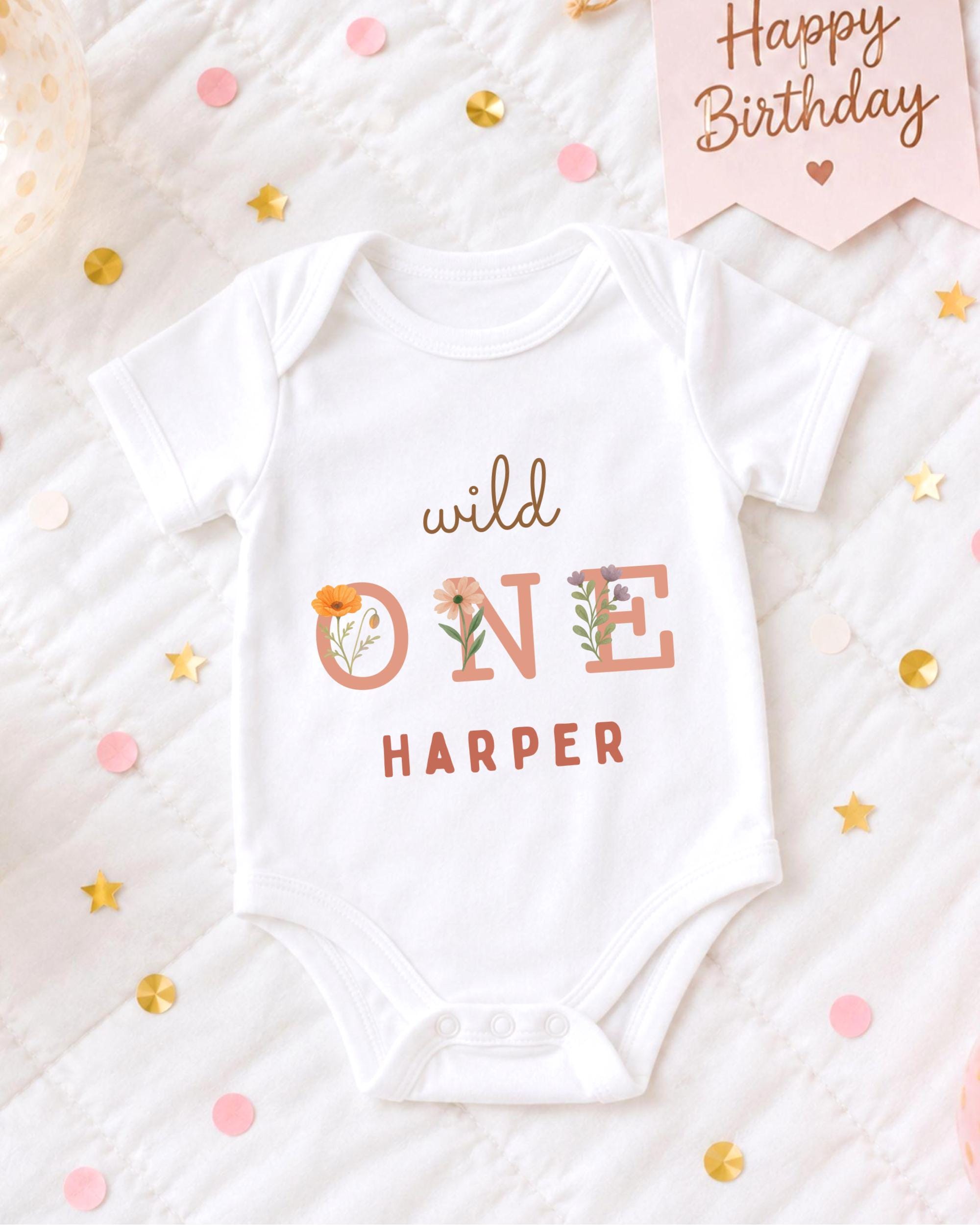 A white onesie with the text "wild one Harper" printed on it, surrounded by confetti and a birthday card.