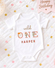 A white onesie with the text "wild one Harper" printed on it, surrounded by confetti and a birthday card.