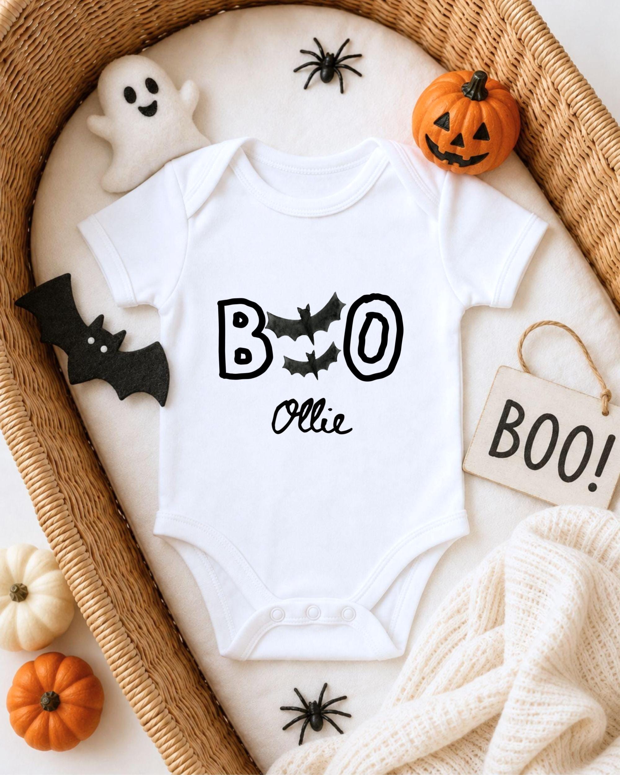 A white onesie with the text "BOO Olle" printed on it, surrounded by Halloween-themed decorations such as a ghost, a pumpkin, and bats.