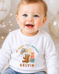 A smiling baby boy wearing a white shirt with a birthday message and a teddy bear graphic.