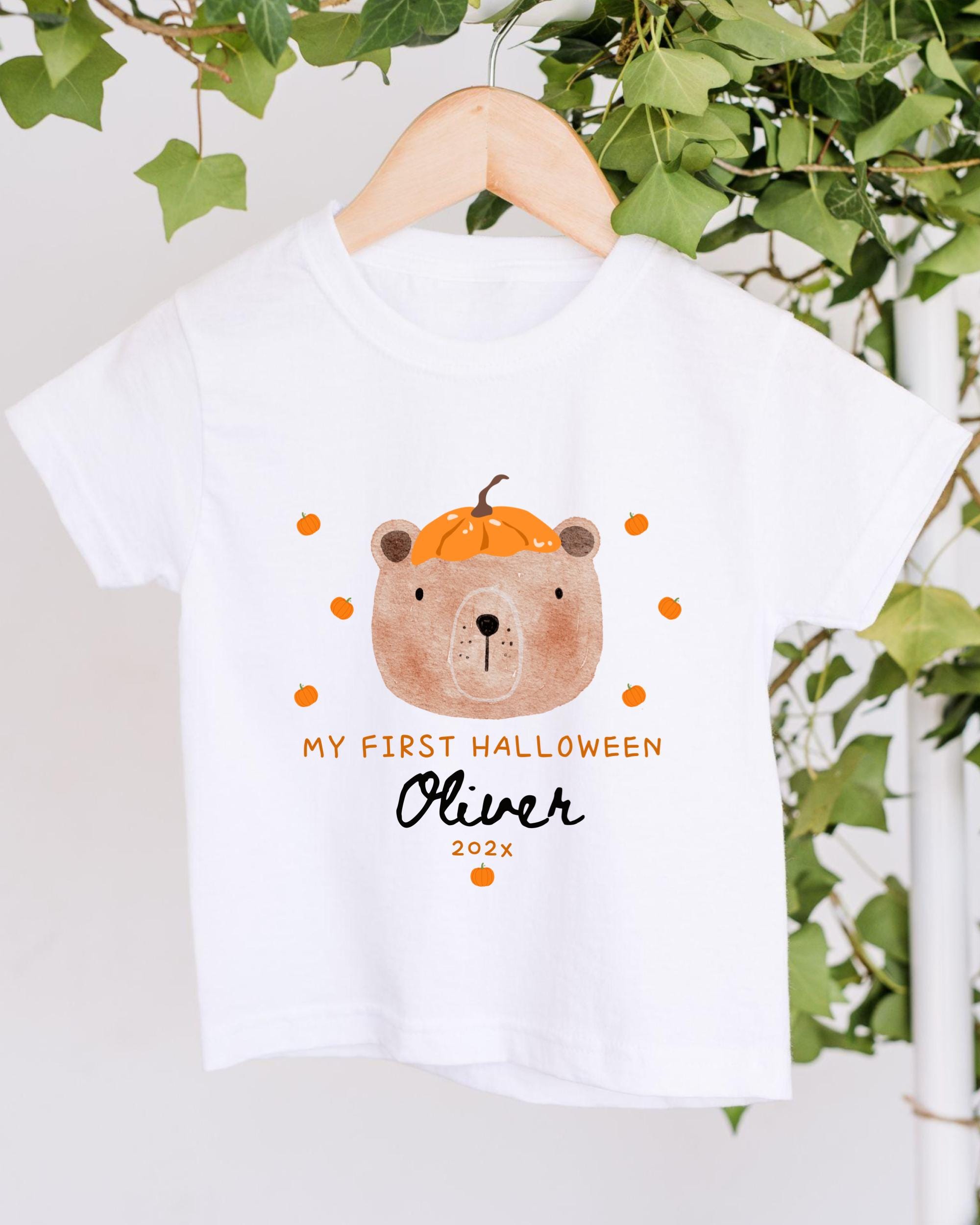 A white t-shirt with a cartoon bear wearing a pumpkin hat and surrounded by orange pumpkins, along with the text "My first Halloween Oliver 2020".