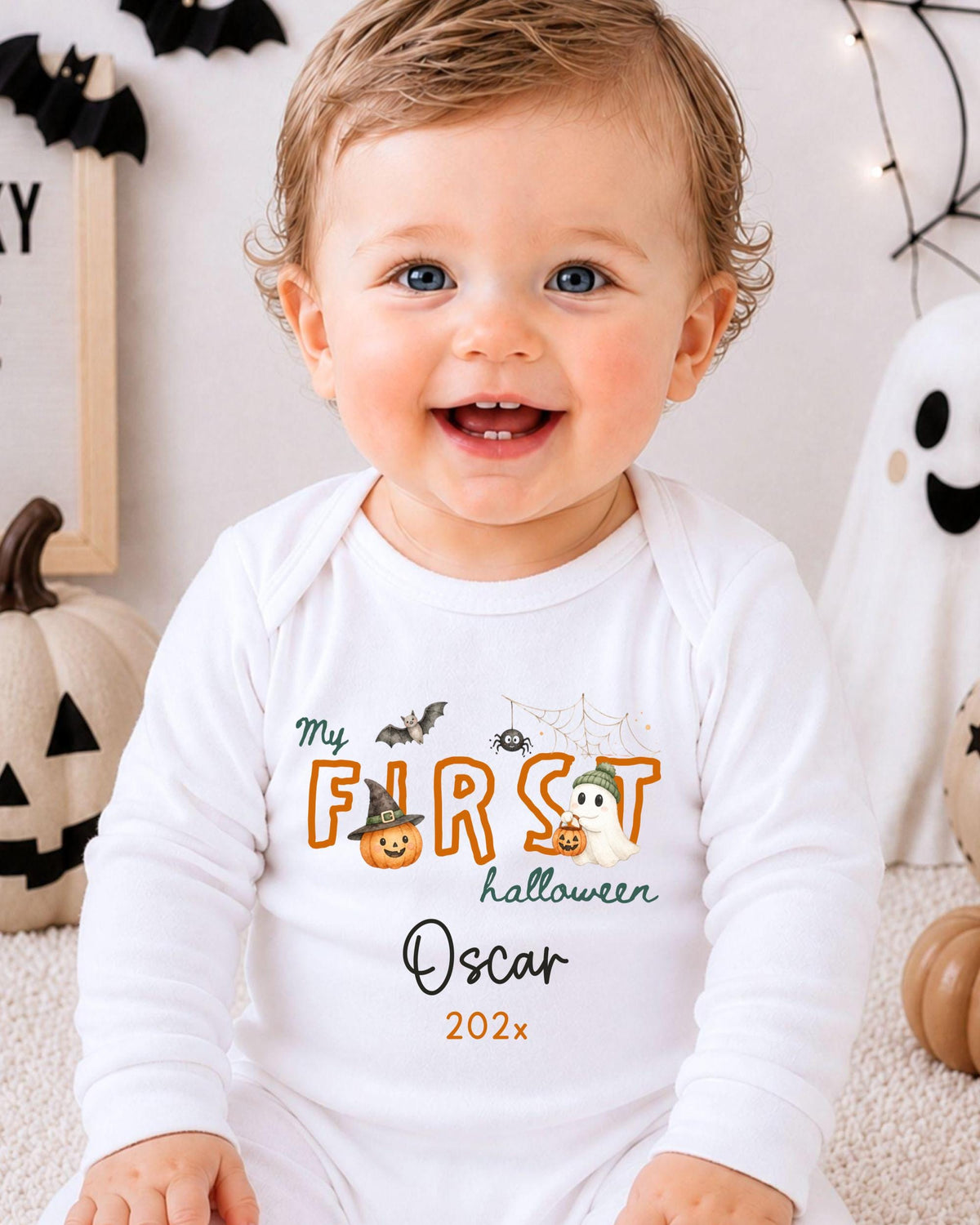 A smiling baby boy wearing a white onesie with a Halloween-themed design, sitting in front of Halloween decorations.