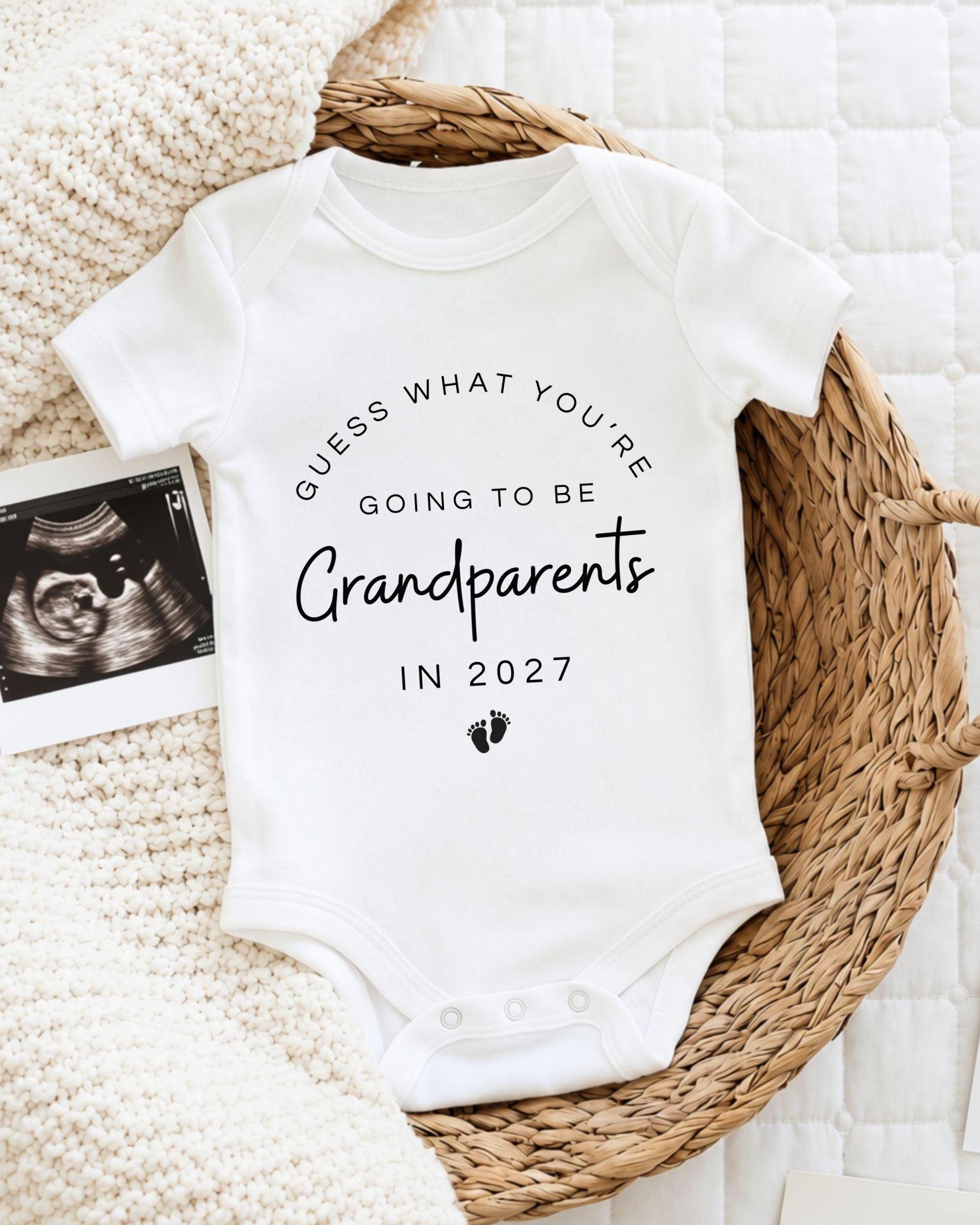 A white baby onesie with text that says "Guess what you're going to be Grandparents in 2027" is displayed on a woven basket.