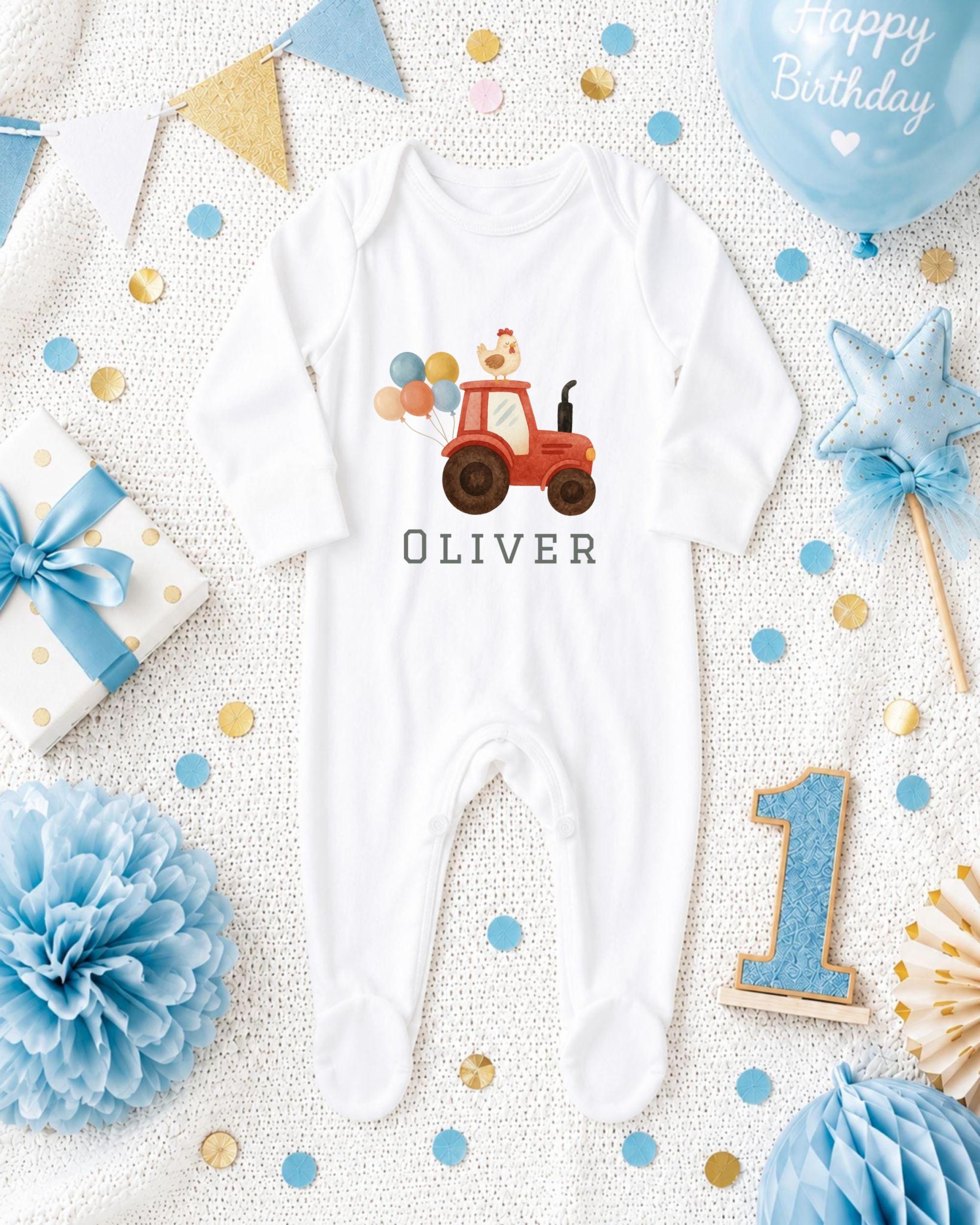 A white onesie with a tractor design, surrounded by birthday decorations such as a blue balloon, blue and gold star, blue and white polka dot gift box, blue and white pom pom, and blue and white confetti.