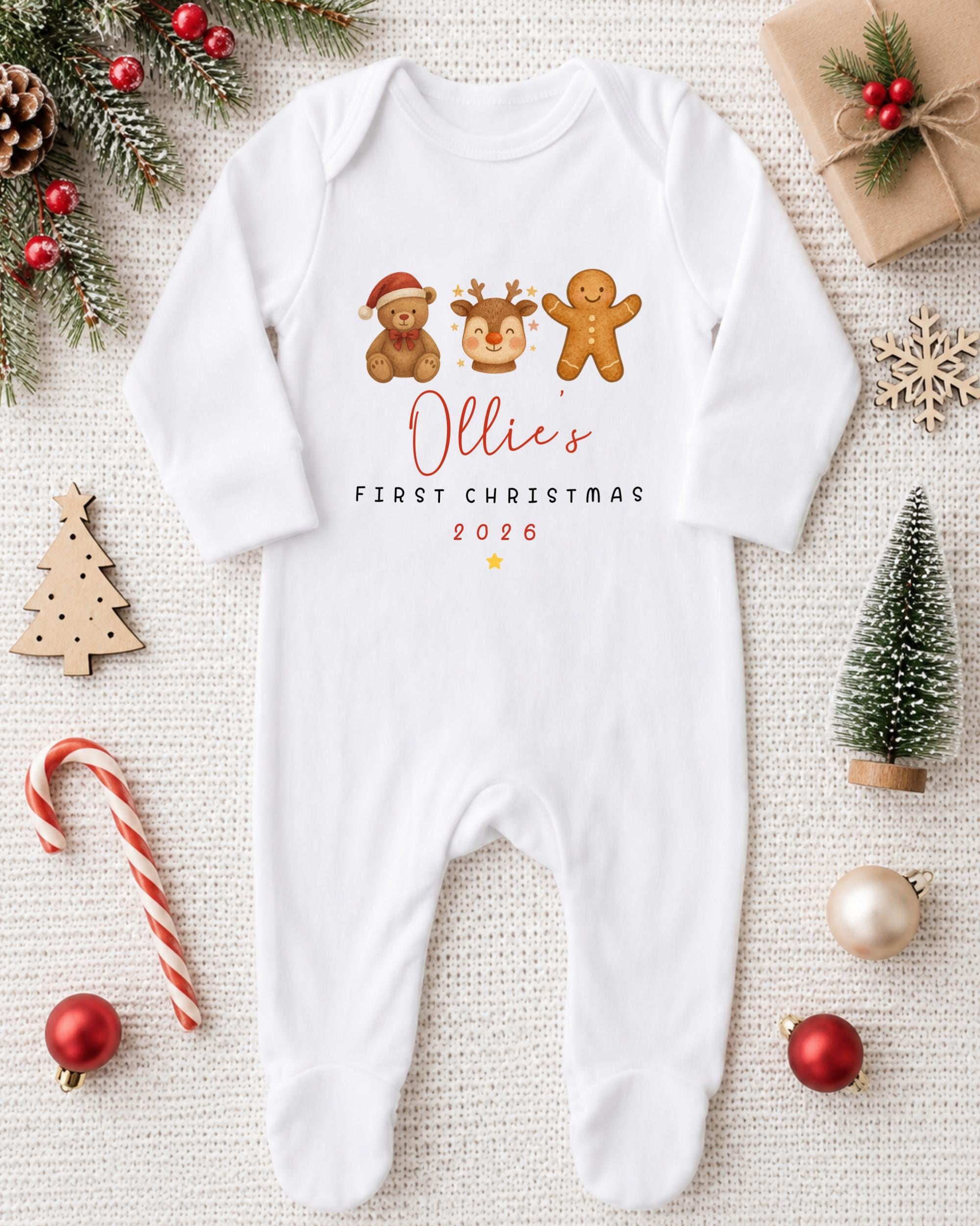 A white onesie with a personalized Christmas design, featuring a teddy bear, gingerbread man, and reindeer, along with the text "Ollie's FIRST CHRISTMAS 2016". The onesie is surrounded by various Christmas decorations, including a pine tree, ornaments, and a candy cane.