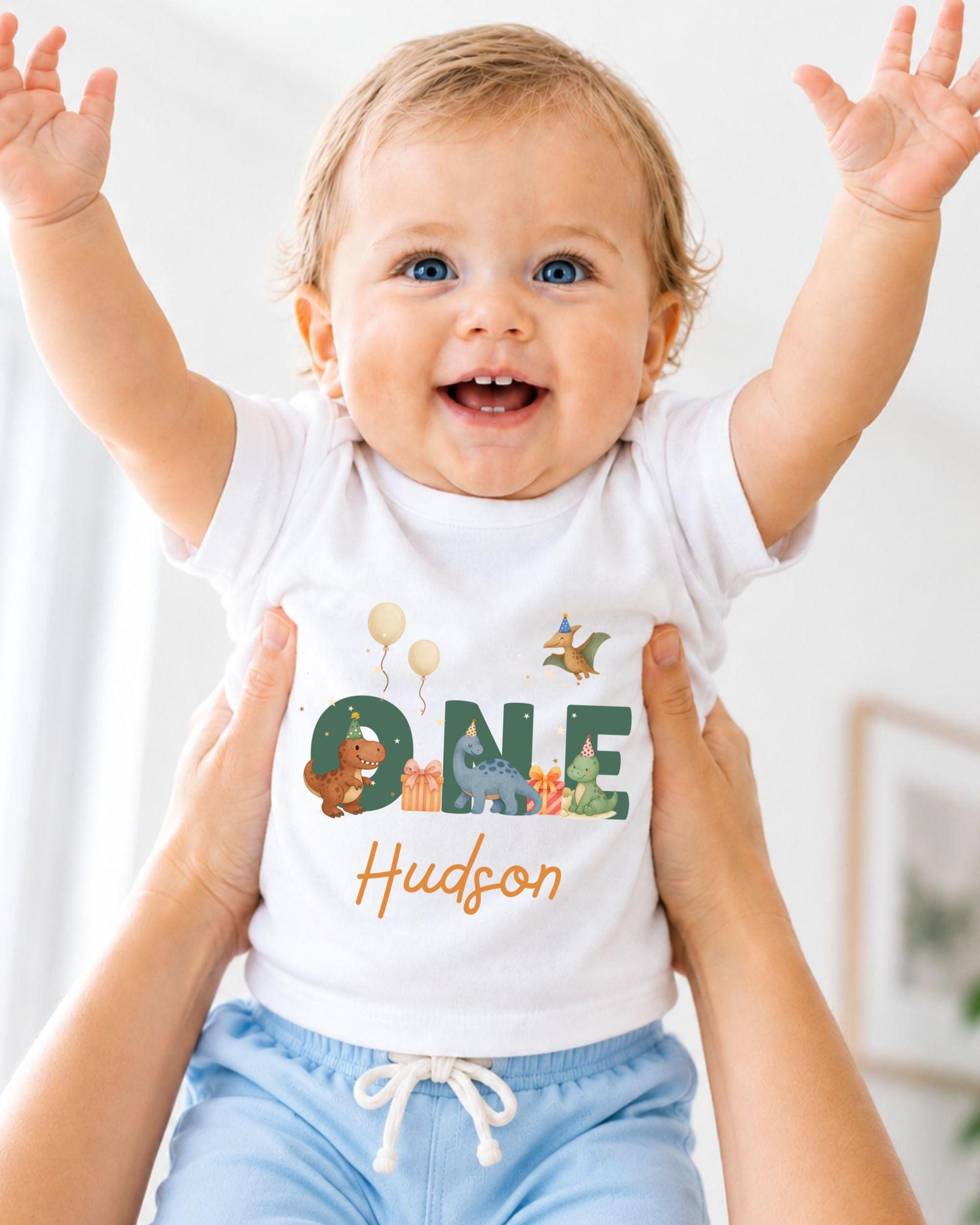 A smiling baby wearing a white t-shirt with a dinosaur and birthday-themed design, being held up by someone's arms.