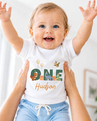 A smiling baby wearing a white t-shirt with a dinosaur and birthday-themed design, being held up by someone's arms.
