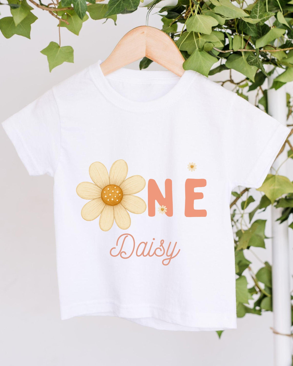 A white t-shirt with a yellow flower design and the text "Daisy" printed on it, hanging on a wooden hanger.
