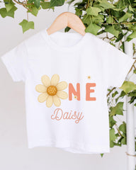 A white t-shirt with a yellow flower design and the text "Daisy" printed on it, hanging on a wooden hanger.