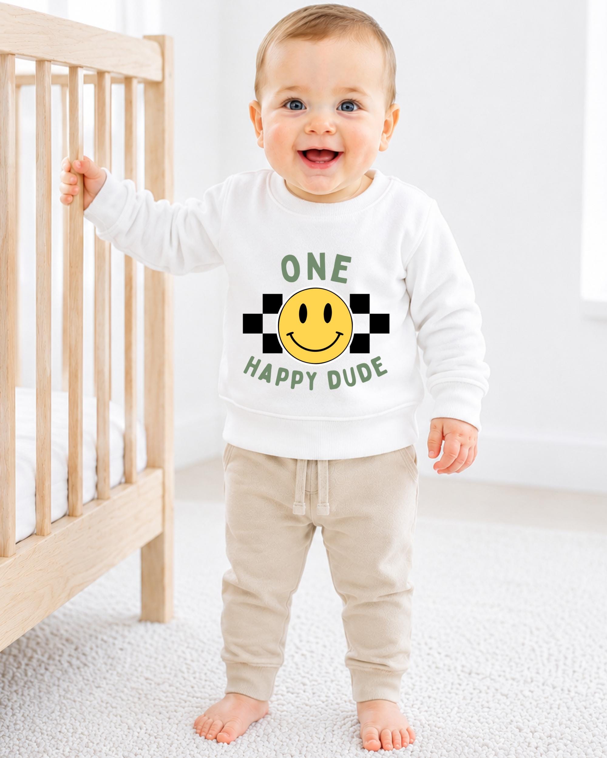 A smiling baby boy wearing a white sweatshirt with a smiley face and the text "ONE HAPPY Dude" is standing next to a wooden crib.