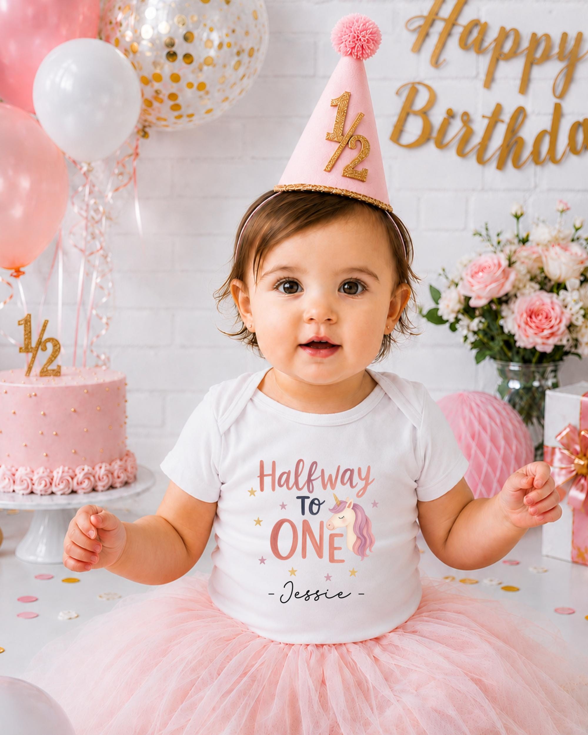 A young girl wearing a birthday hat and a white t-shirt with a unicorn design is sitting in front of a birthday cake and balloons.