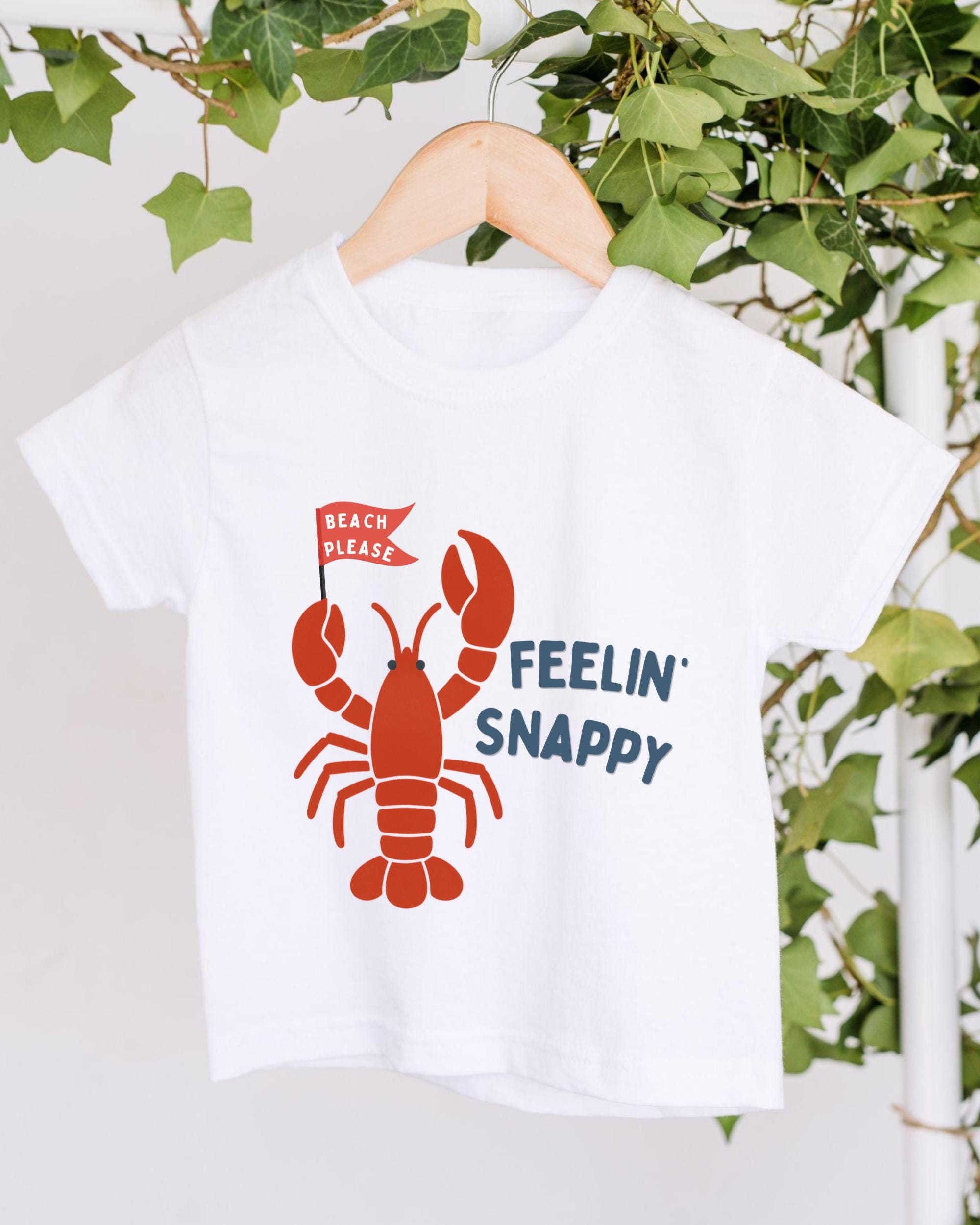 A white t-shirt with a red lobster graphic and text printed on it, hanging on a wooden hanger.