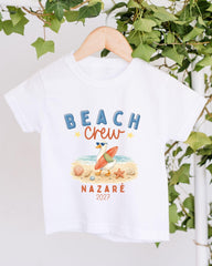A white t-shirt with a beach-themed graphic and text printed on it, hanging on a wooden hanger.