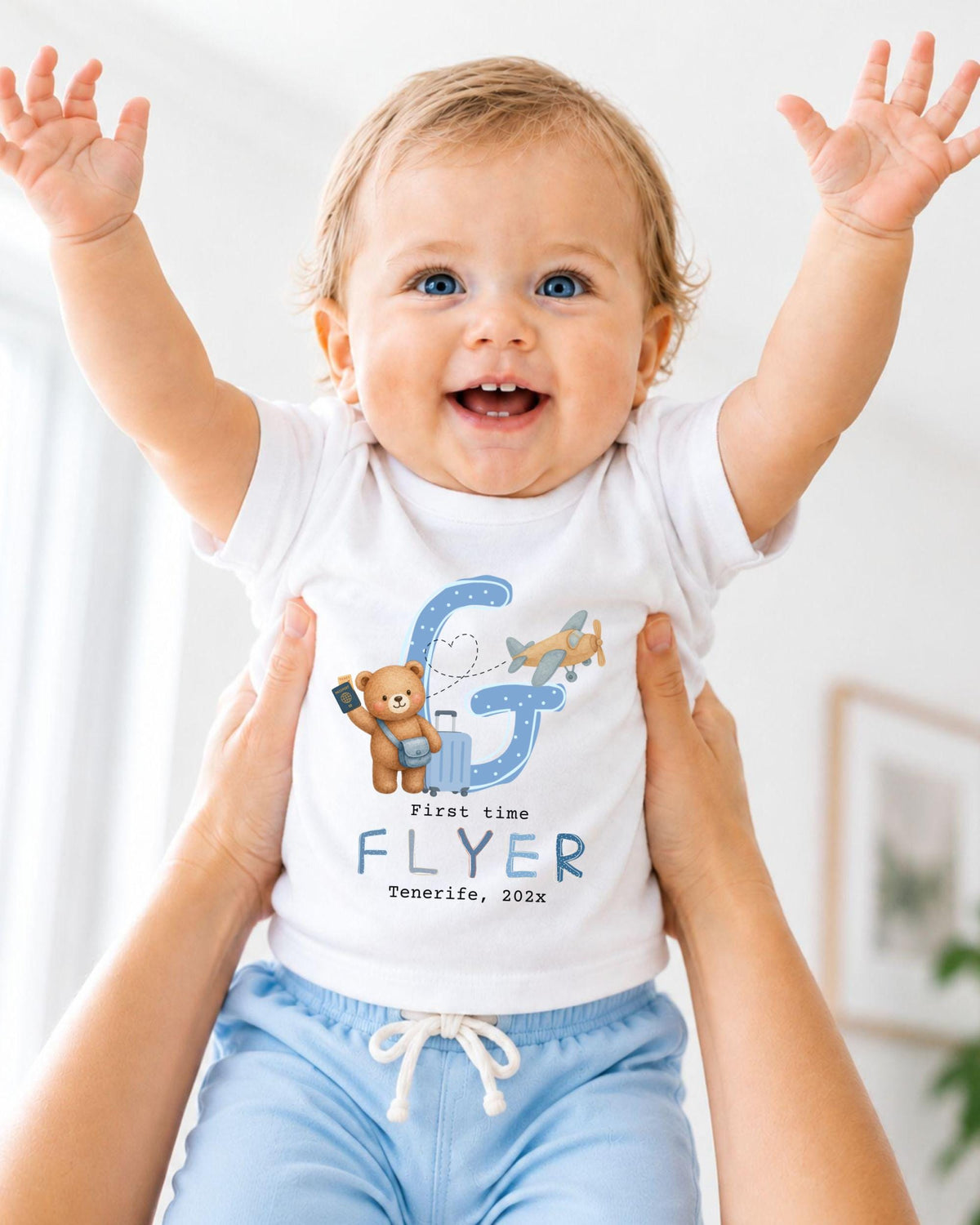 A smiling baby wearing a white shirt with a letter "G" and a teddy bear graphic, being held up by someone's arms.