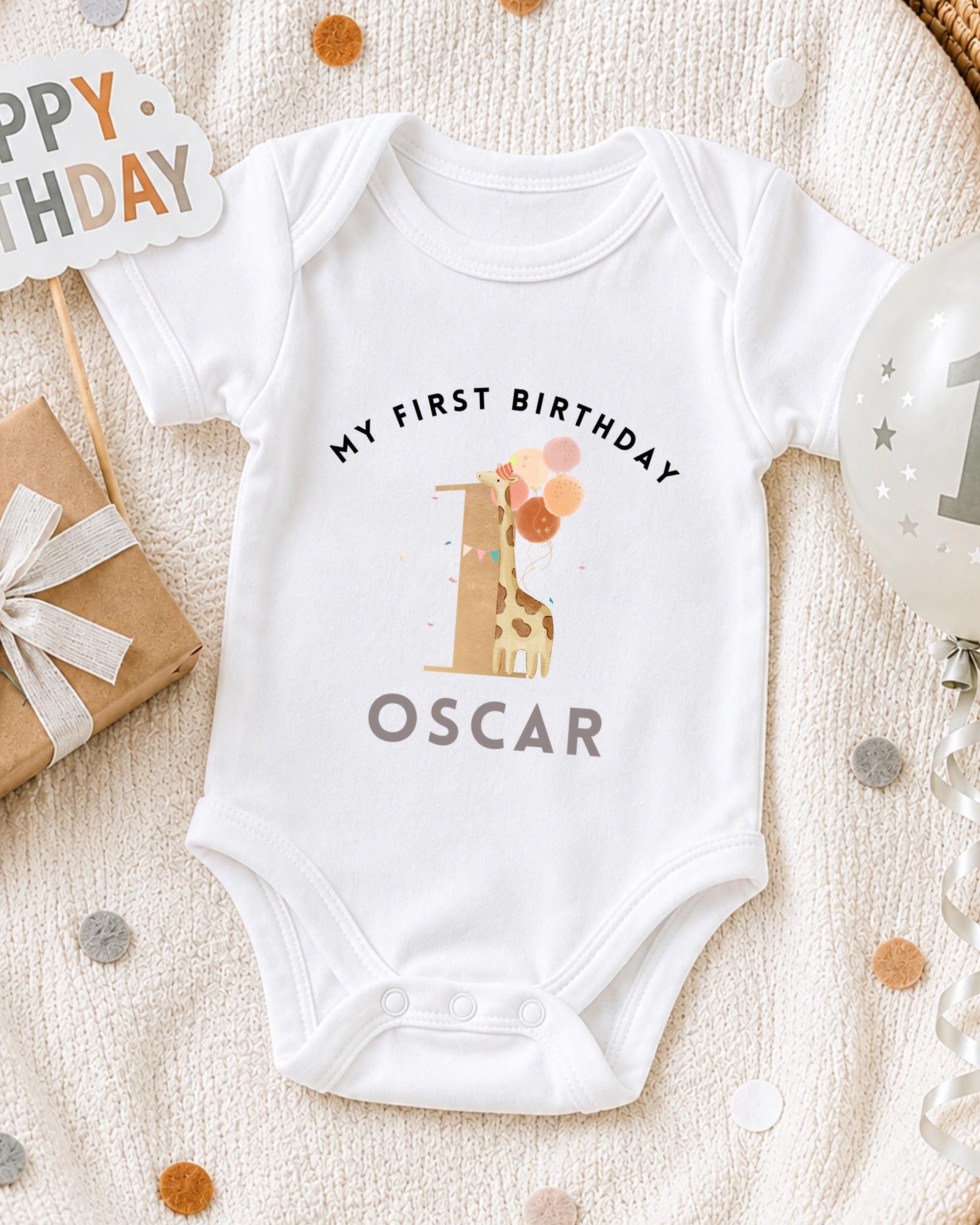 A white onesie with a giraffe and the text "My first birthday Oscar" printed on it, along with a "Happy Birthday" sign and a birthday balloon.