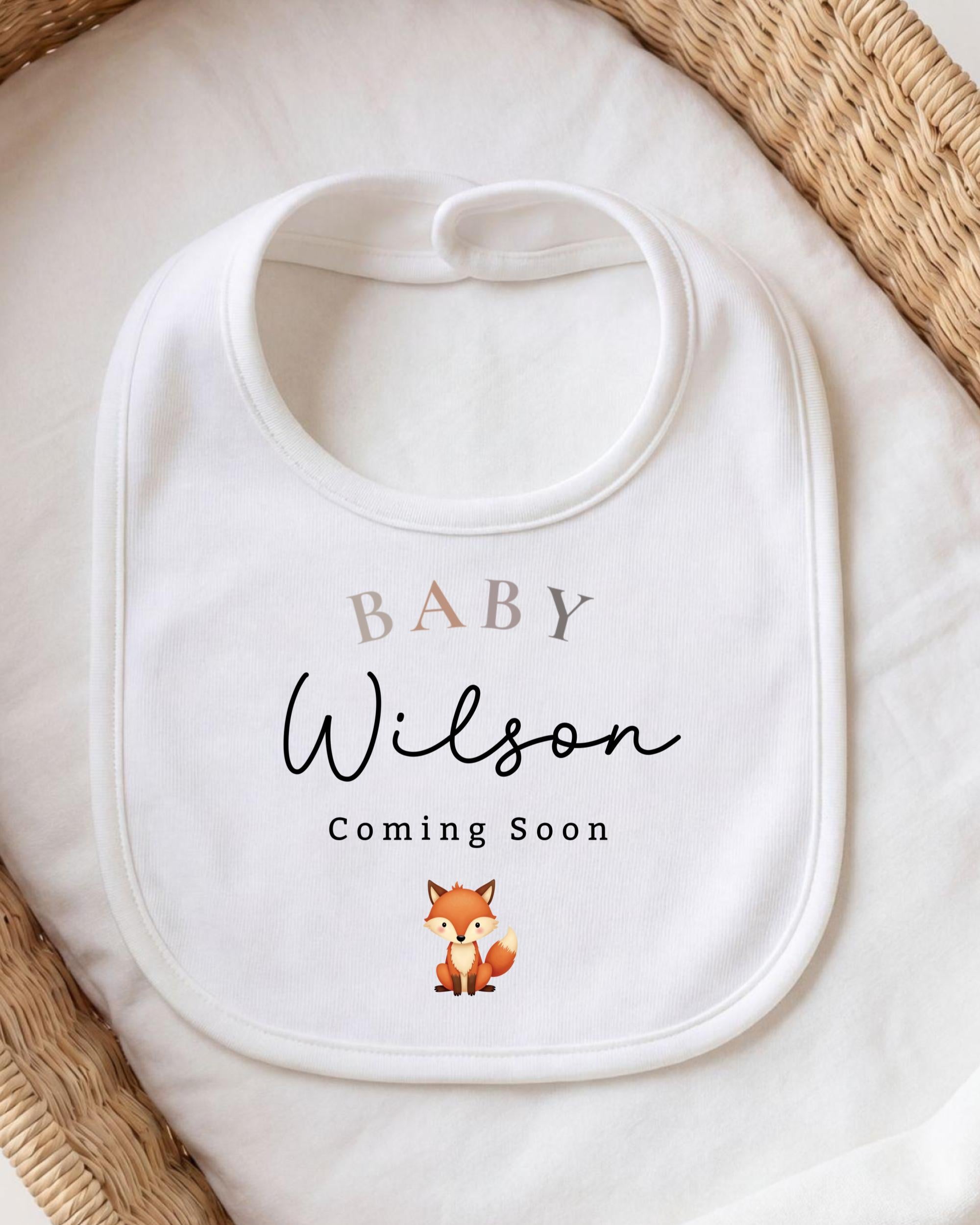 A white baby bib with the text "BABY Wilson Coming Soon" printed on it, along with a small illustration of a fox.