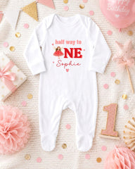 A white onesie with the text "half way to 1E Sophie" printed on it, surrounded by pink and gold confetti, a pink star, and other baby-related items.