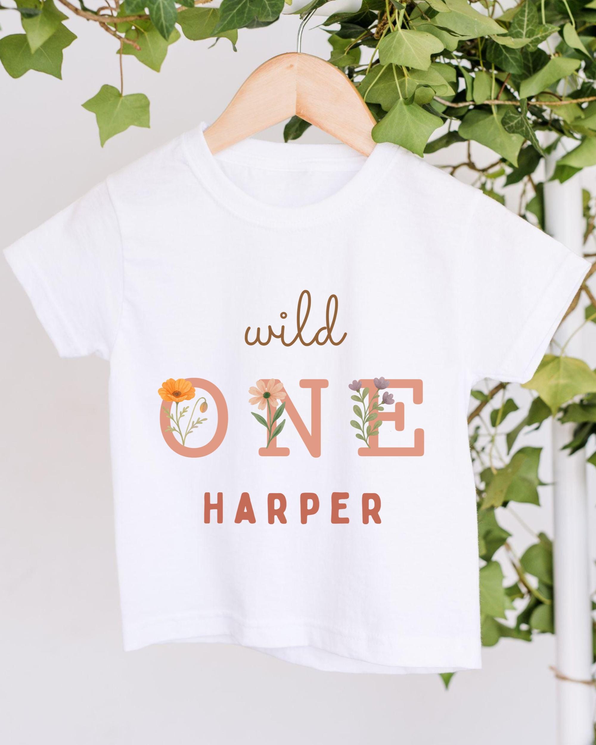A white t-shirt with the text "wild one Harper" printed on it, hanging on a wooden hanger.