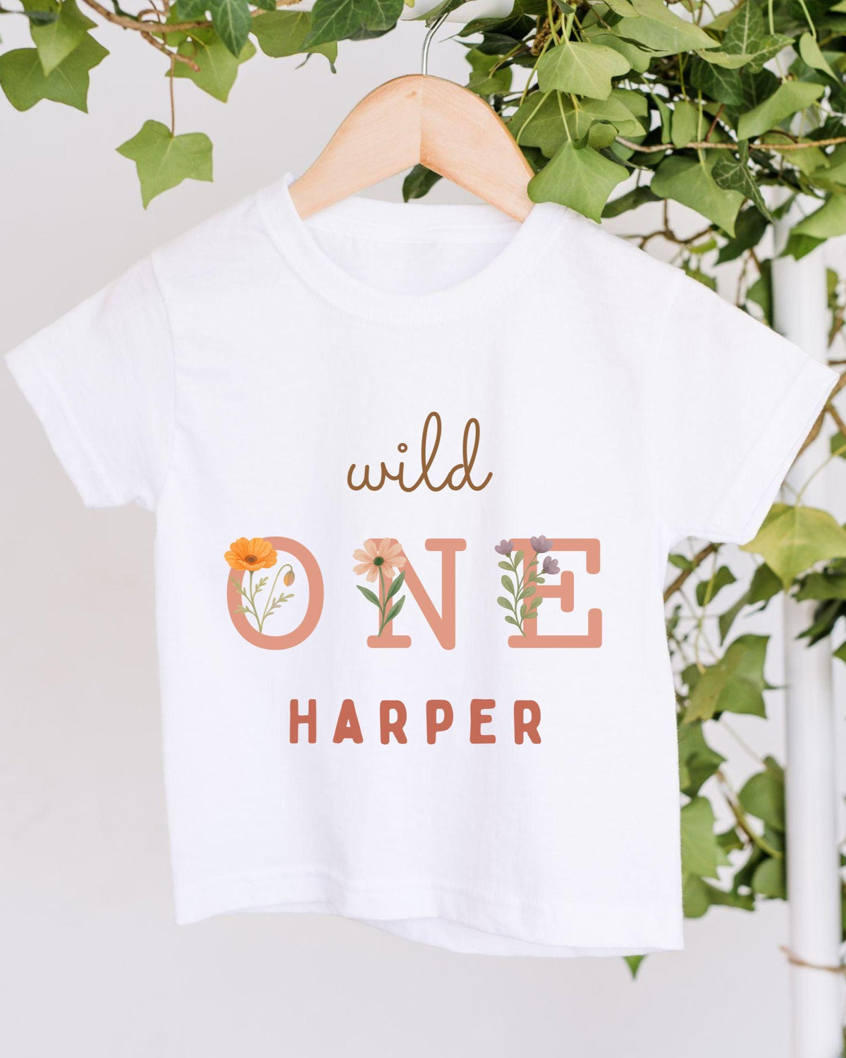 A white t-shirt with the text "wild one Harper" printed on it, hanging on a wooden hanger.