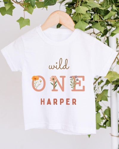 A white t-shirt with the text "wild one Harper" printed on it, hanging on a wooden hanger.