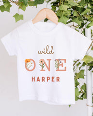 A white t-shirt with the text "wild one Harper" printed on it, hanging on a wooden hanger.