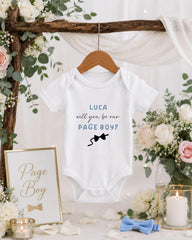 A white baby onesie with the text "LUCA will you be our PAGE BOY?" printed on it, hanging on a wooden hanger. The onesie is displayed in a setting with floral decorations, including a framed sign that reads "Page Boy".