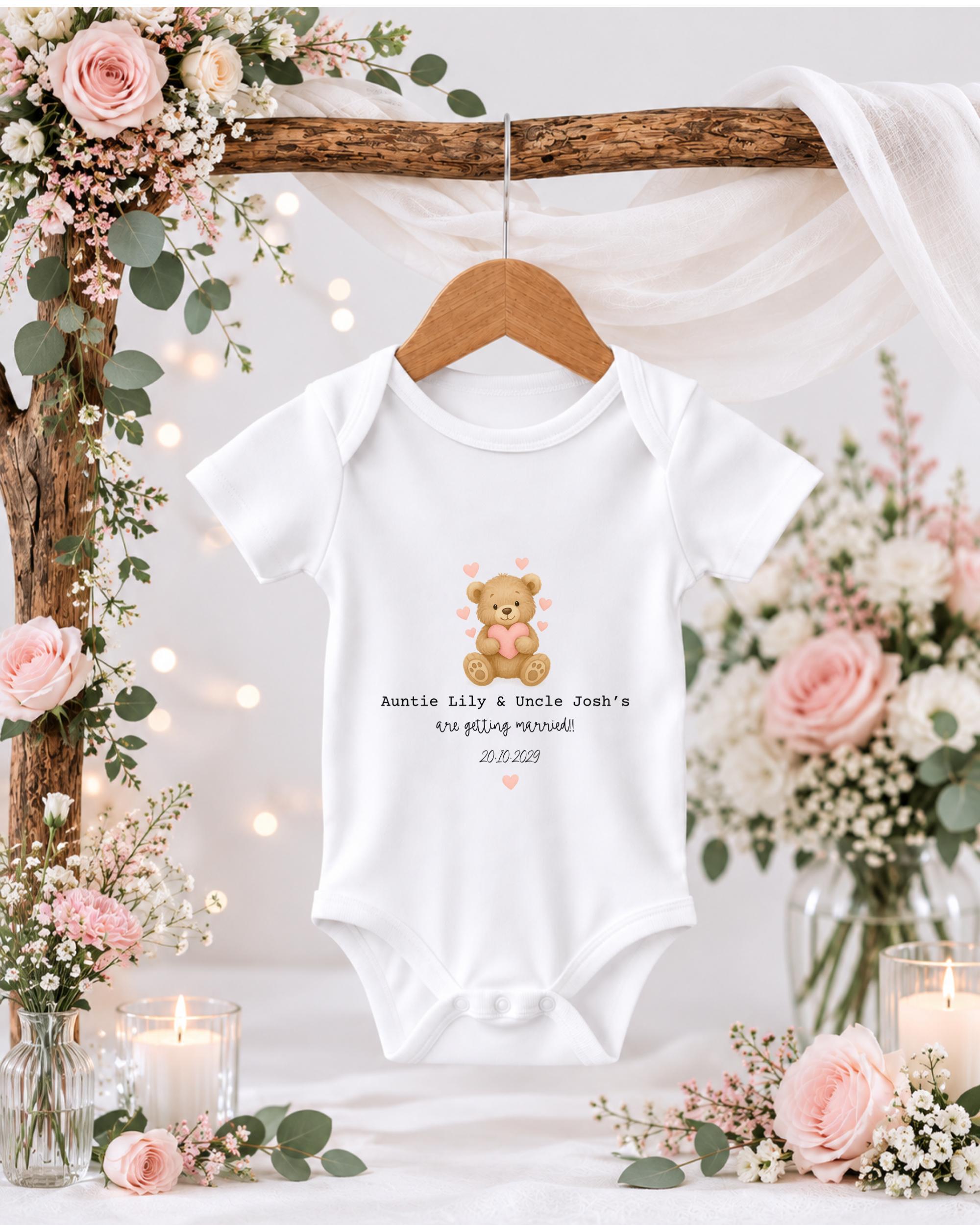 A white baby onesie with a teddy bear graphic hangs on a wooden hanger, surrounded by pink and white flowers and candles.