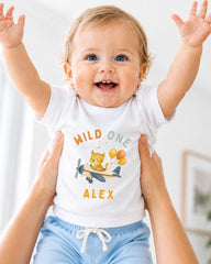 A smiling baby wearing a white t-shirt with a cartoon airplane and the name "Alex" printed on it, being held up by someone's arms.