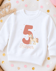 A white sweatshirt with a unicorn and the number 5 printed on it, along with the name "Harper" below the number.