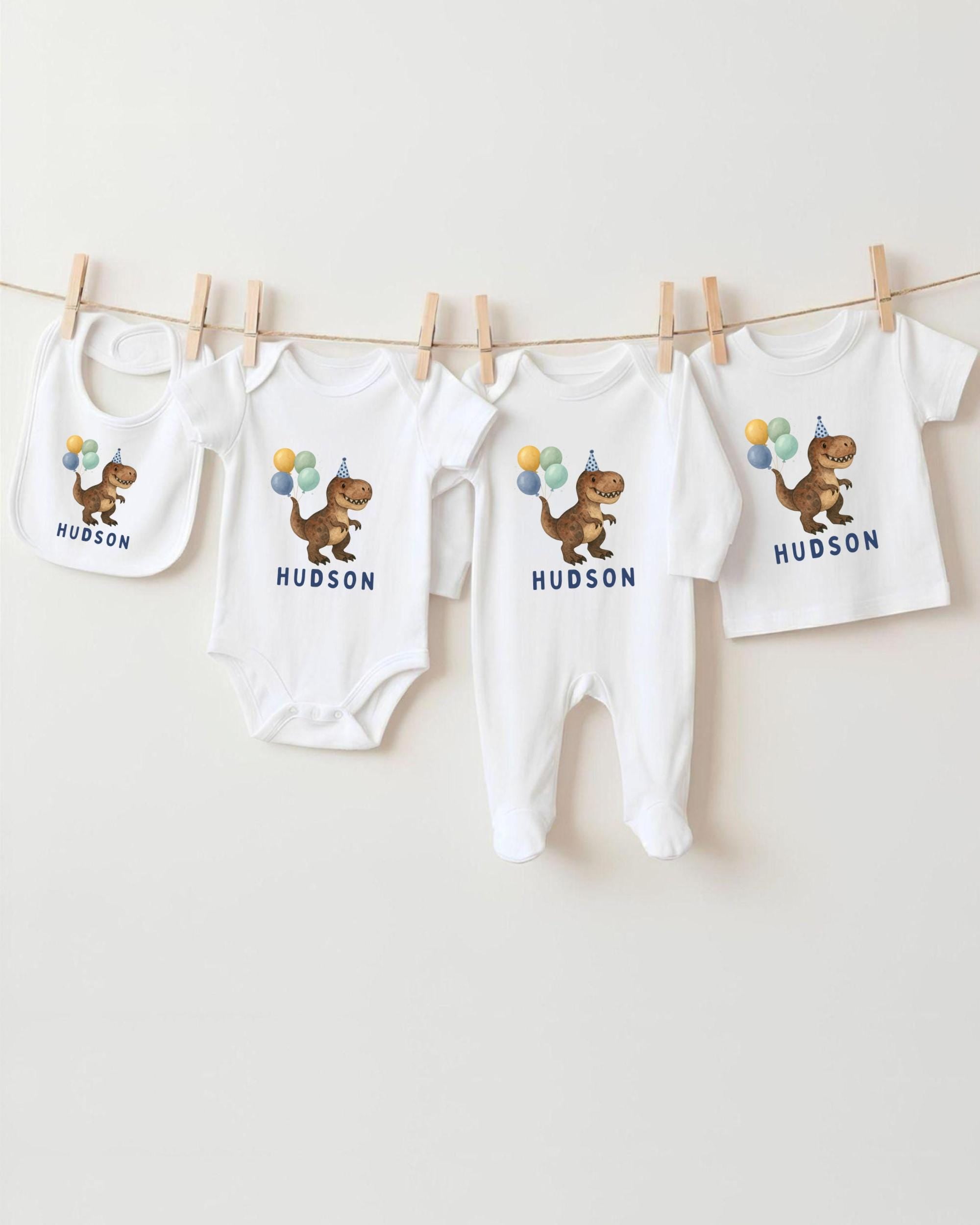 A collection of white baby clothes, including onesies and bibs, with dinosaur illustrations and birthday party balloons hanging on a clothesline.