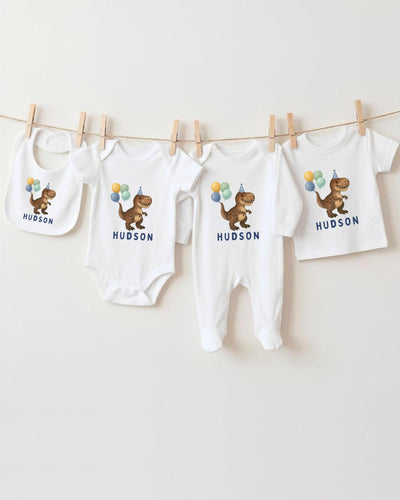 A collection of white baby clothes, including onesies and bibs, with dinosaur illustrations and birthday party balloons hanging on a clothesline.