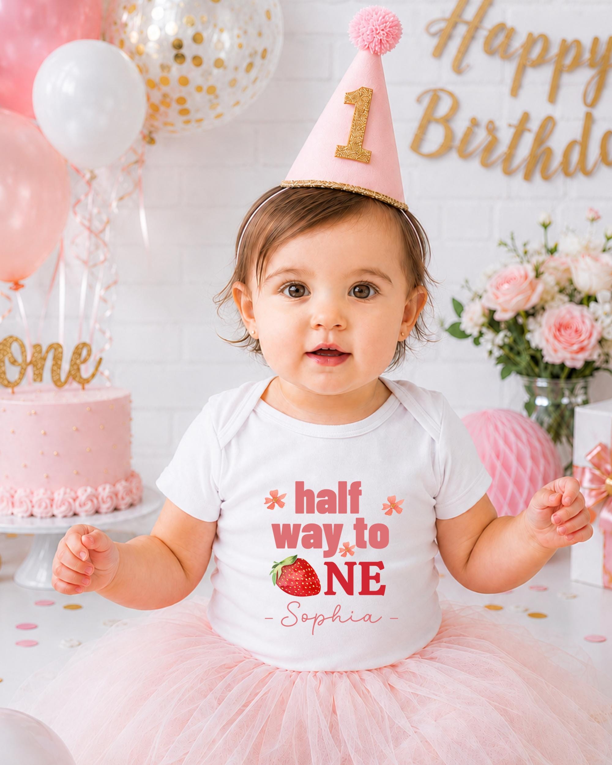 A young girl wearing a birthday hat and a white t-shirt with the text "half way to one" and "Sophia" is posing in front of a birthday cake and balloons.