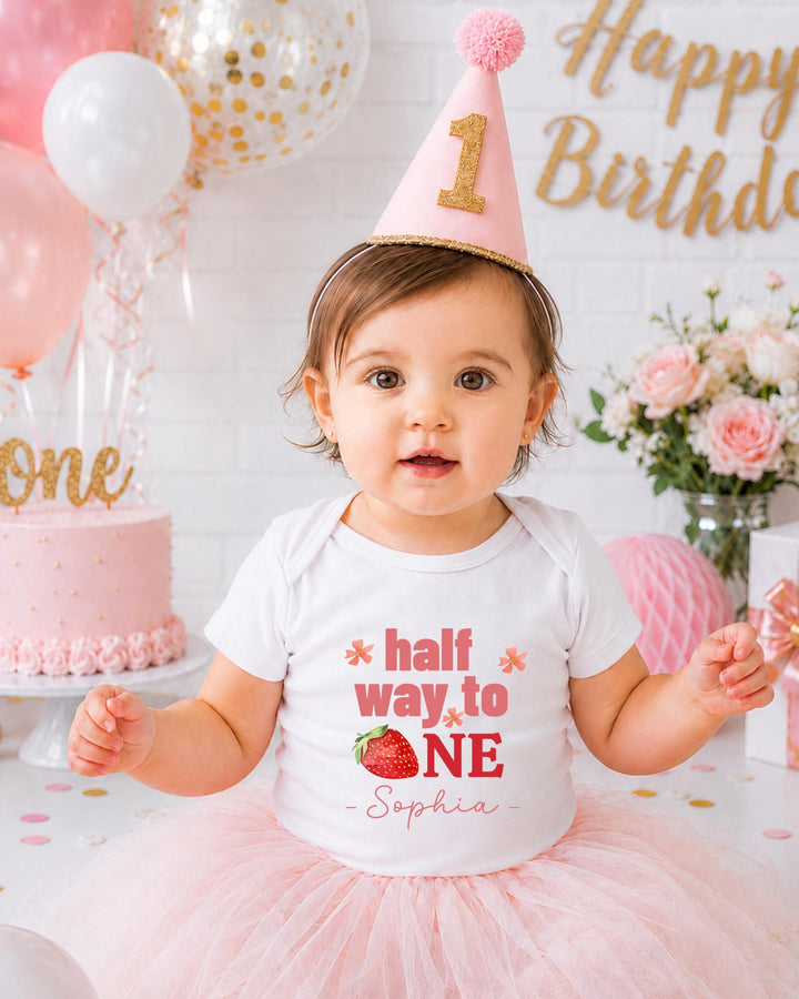 A young girl wearing a birthday hat and a white t-shirt with the text "half way to one" and "Sophia" is posing in front of a birthday cake and balloons.