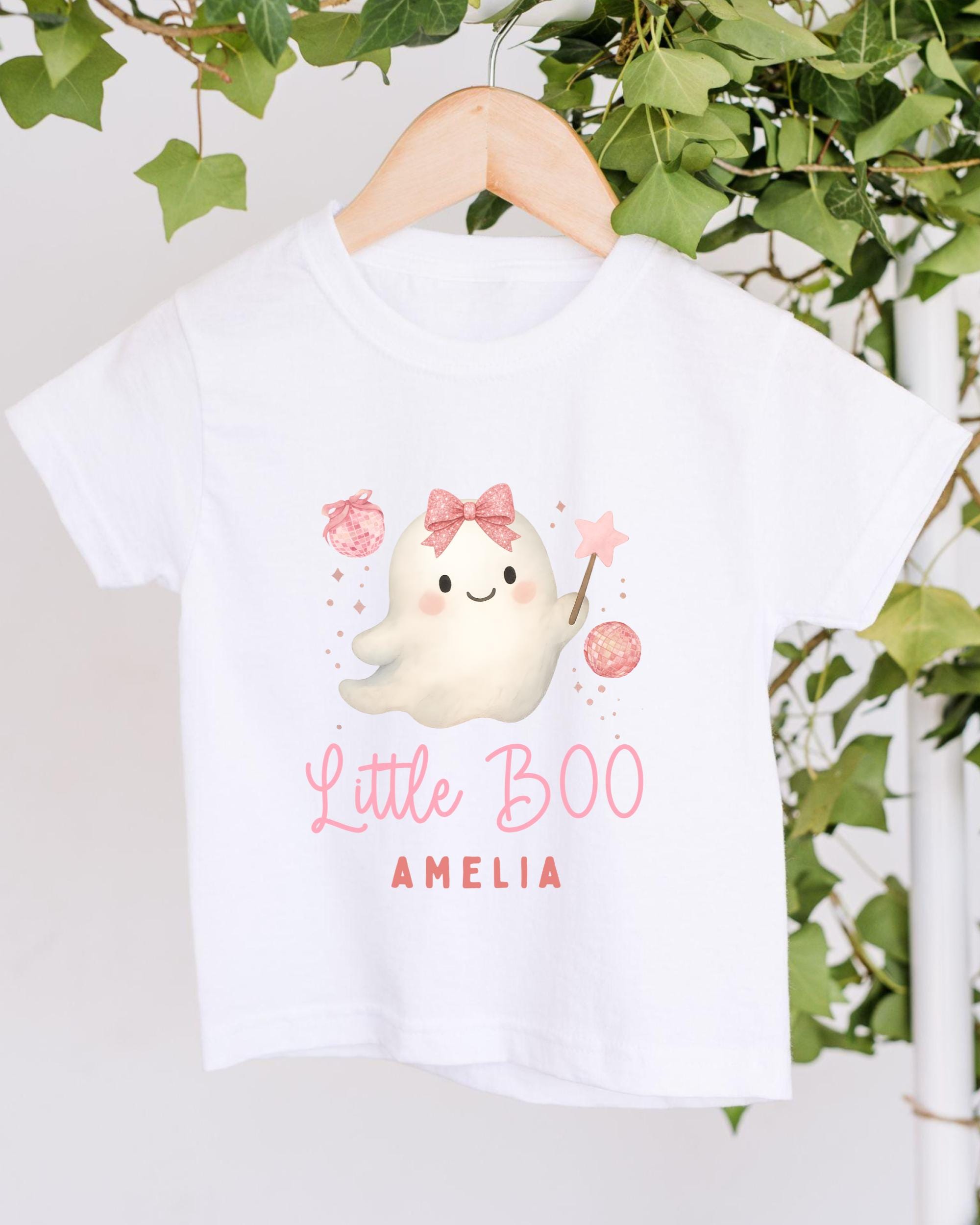 A white t-shirt with a cartoon ghost character and text printed on it, hanging on a wooden hanger.