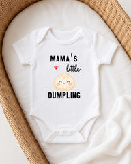 A white onesie with a cartoon image of a dumpling and the text "Mama's little DUMPLING" printed on it, laid out on a white cloth.