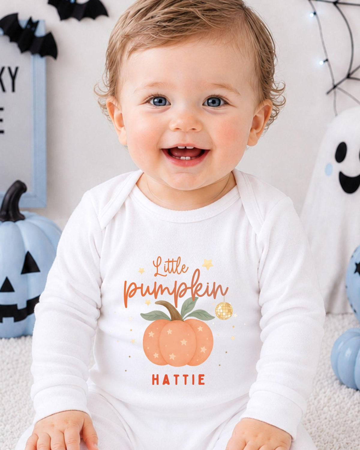 A smiling baby boy wearing a white onesie with a pumpkin and stars on it, sitting in front of Halloween decorations.