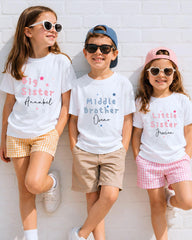 Three children wearing white t-shirts with personalized text, smiling and posing against a white brick wall.