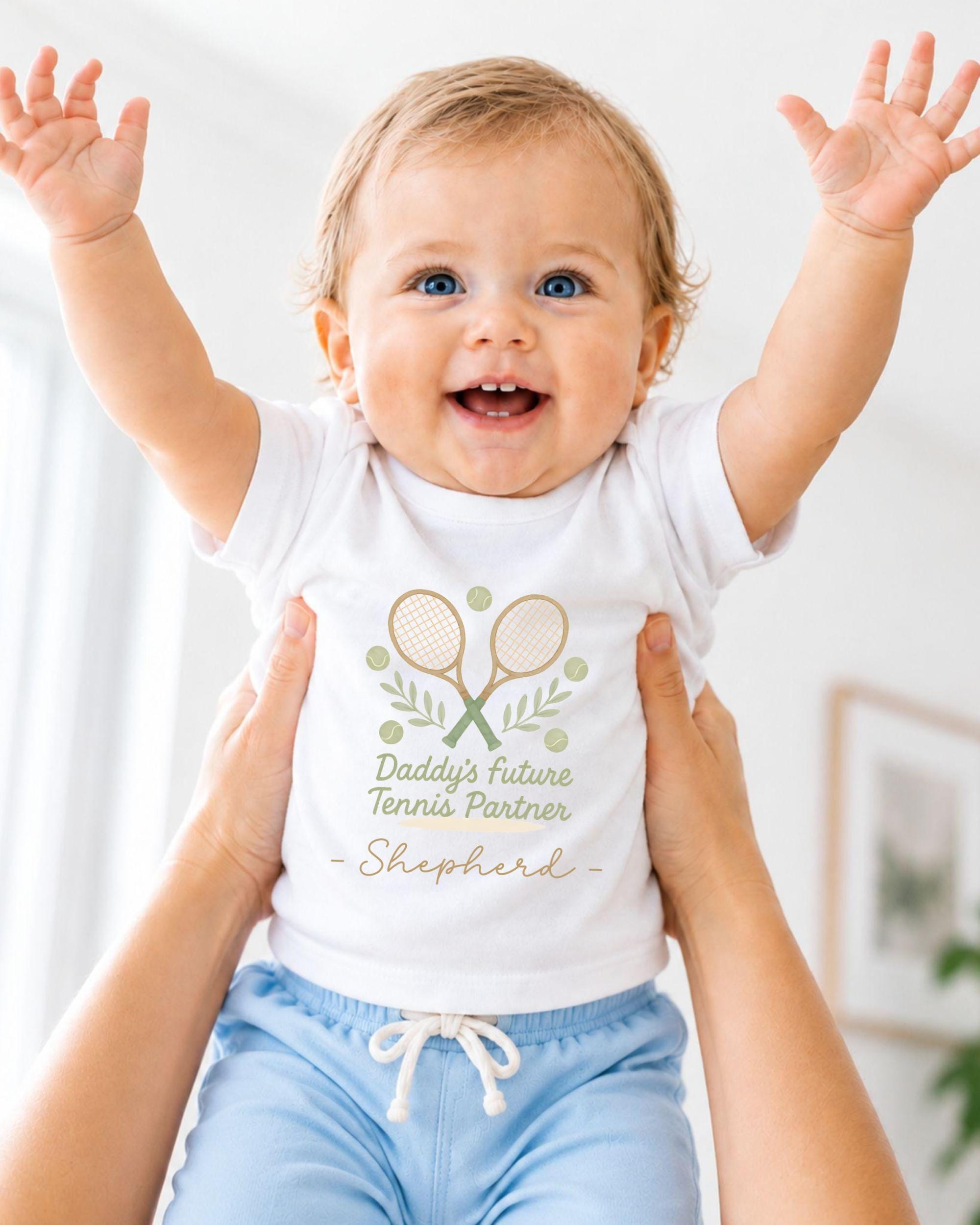 A smiling baby wearing a white t-shirt with a tennis racket and leaves design, being held up by someone's arms.