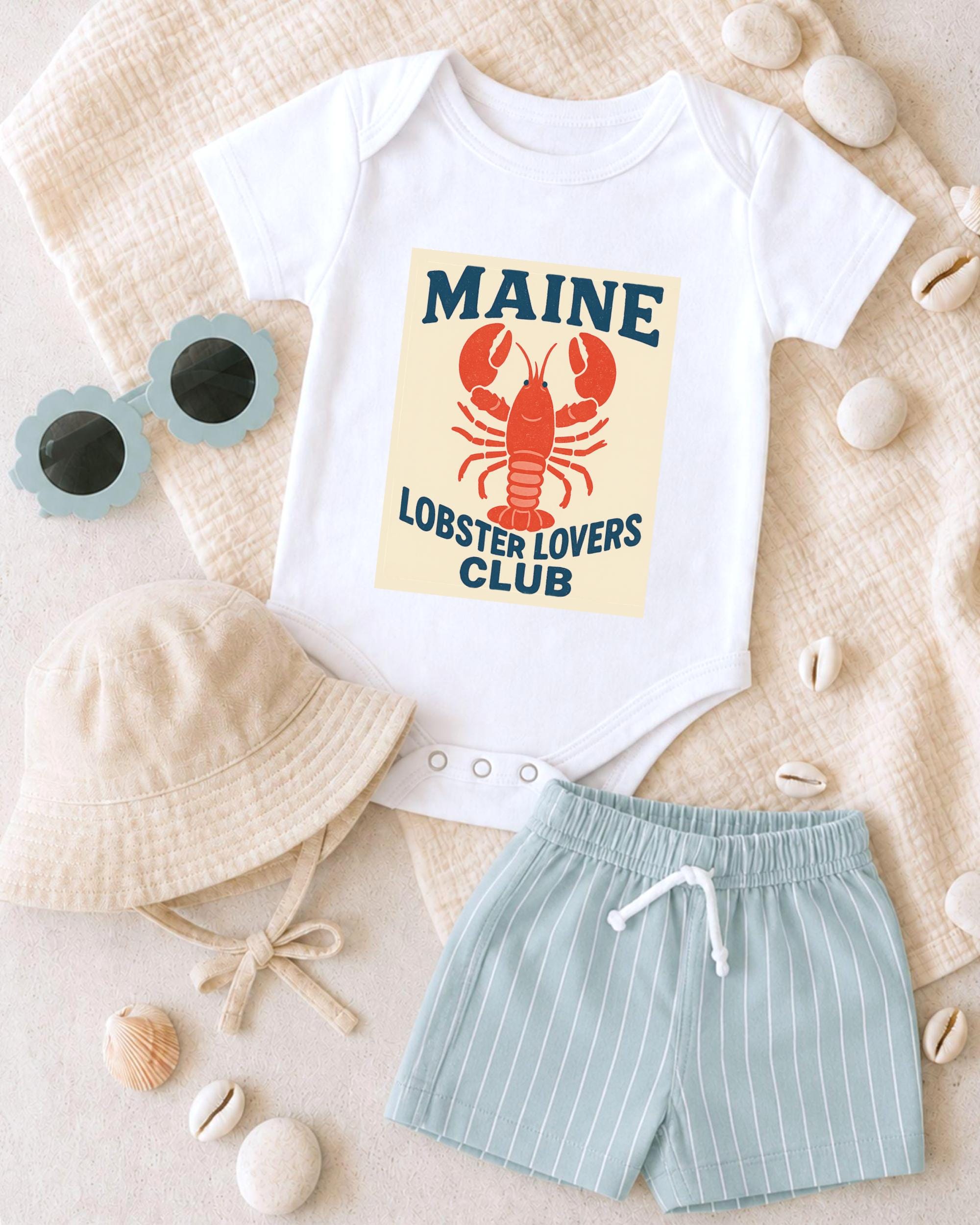 A white onesie with a lobster design, a beige bucket hat, and light blue shorts are arranged on a beige surface, along with sunglasses and seashells.
