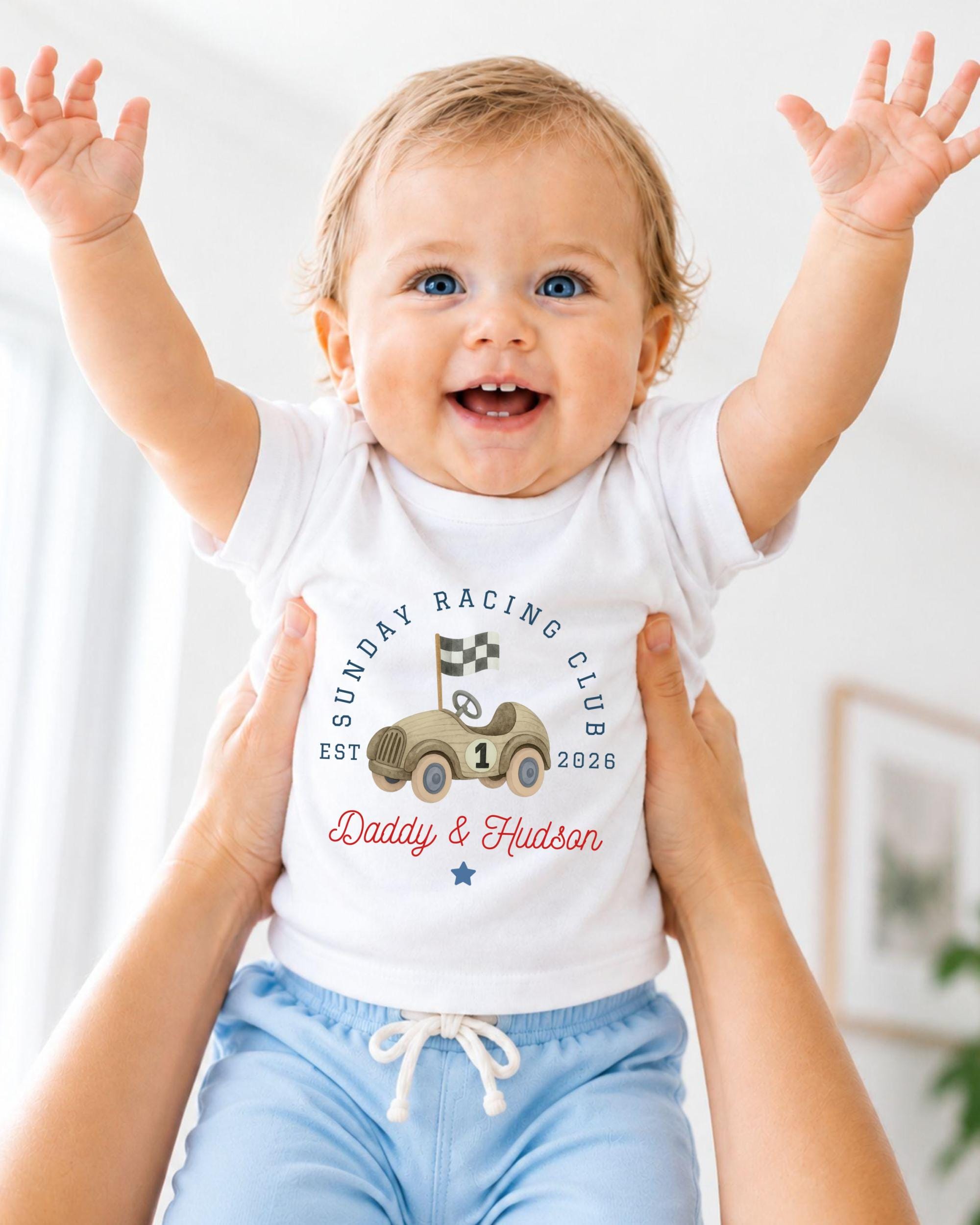 A smiling baby wearing a white t-shirt with a graphic of a race car and the text "Sunday Racing Club EST. 1926" is being held up by two people.