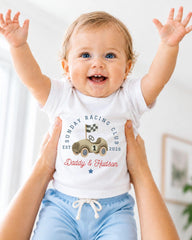 A smiling baby wearing a white t-shirt with a graphic of a race car and the text "Sunday Racing Club EST. 1926" is being held up by two people.