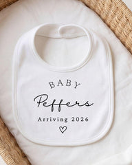 A white baby bib with the text "BABY Peppers Arriving 2026" printed on it, placed on a white cloth.