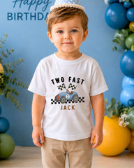A young boy wearing a white t-shirt with a racing car graphic and the text "Two Fast Jack" stands in front of a birthday party backdrop with balloons.