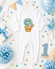 A baby onesie with a hot air balloon and a lion on it, surrounded by birthday decorations such as a blue balloon, a star, and confetti.